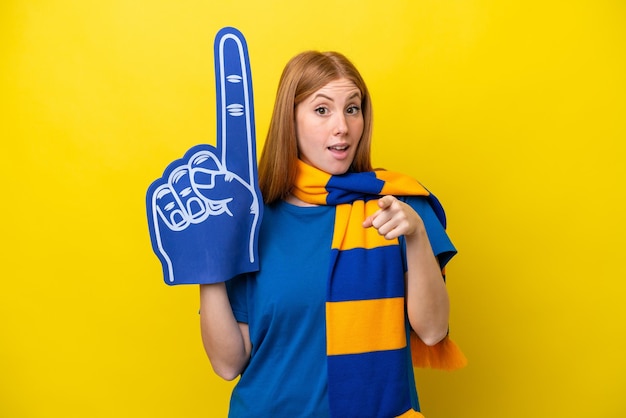 Young redhead sports fan woman isolated on yellow background\
surprised and pointing front