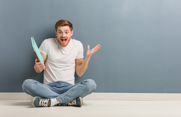 Young redhead student man sitting on the floor celebrating a victory or success. He is holding books.