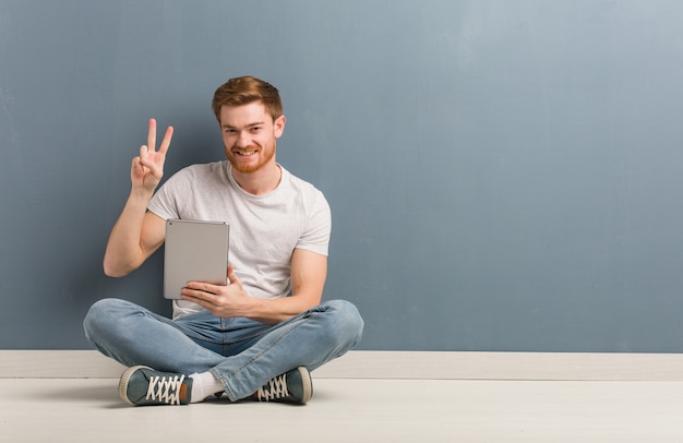 Young redhead student man sitting on the floor fun and happy doing a gesture of victory. He is holding a tablet.