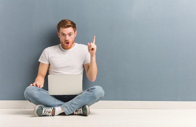 Young redhead student man sitting on the floor having a great idea, concept of creativity. He is holding a laptop.