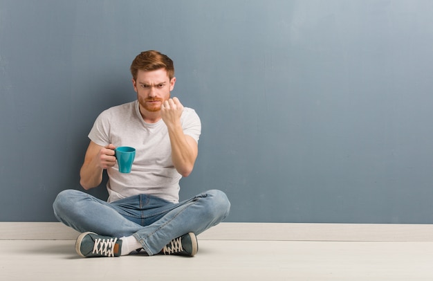 Giovane uomo dello studente della testarossa che si siede sul pavimento che mostra pugno alla fronte, espressione arrabbiata. ha in mano una tazza di caffè.