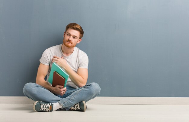 自信を持って、腕を組んで、見上げて床に座っている赤毛の若い学生男。