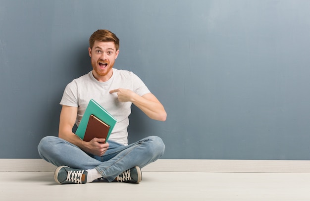 Young redhead student man sitting on the floor surprised, feels successful and prosperous. He is holding books.