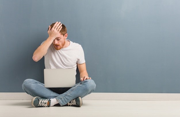 Young redhead student man sitting on the floor worried and overwhelmed. He is holding a laptop.