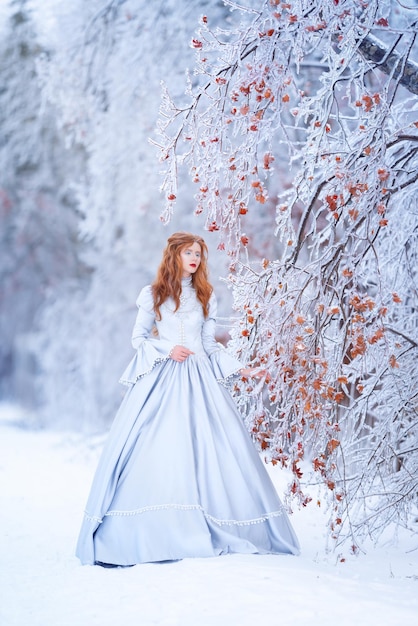 Фото Молодая рыжая принцесса гуляет в зимнем лесу в синем платье мороз и снег