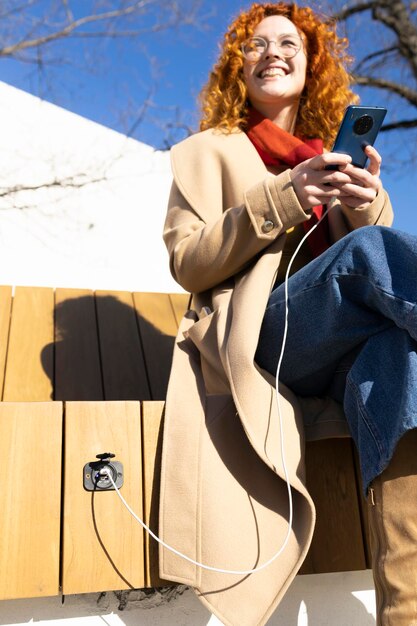 Young redhead woman charging her smartphone on a smart bench on a warm spring day