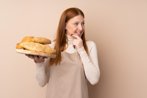 Young redhead woman in chef uniform. Female baker holding a table with several breads thinking an idea and looking side