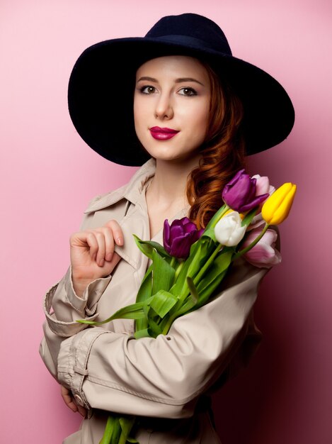 Молодая рыжая женщина в плаще с букетом тюльпанов на розовом
