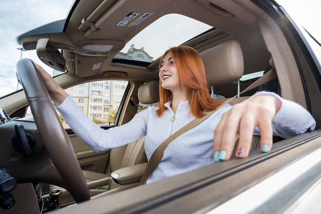 молодая рыжая женщина-водитель пристегнута ремнем безопасности за рулем автомобиля, счастливо улыбаясь
