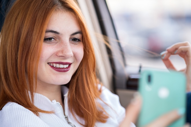 ラッシュアワーの交通渋滞で車のホイールの後ろに座っている彼女の携帯電話でselfiesを取って若い赤毛の女性ドライバー。