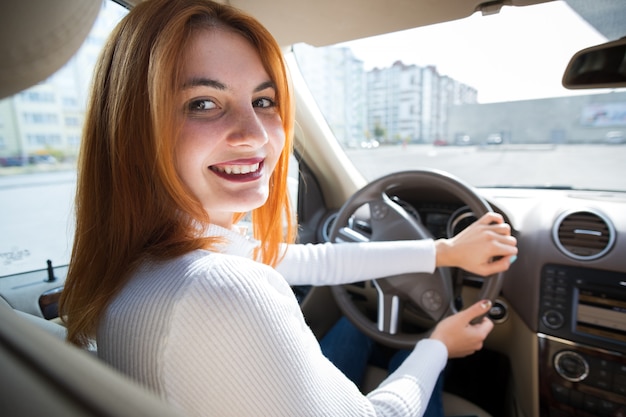 Young redhead woman driver behind a wheel driving a car smiling happily.