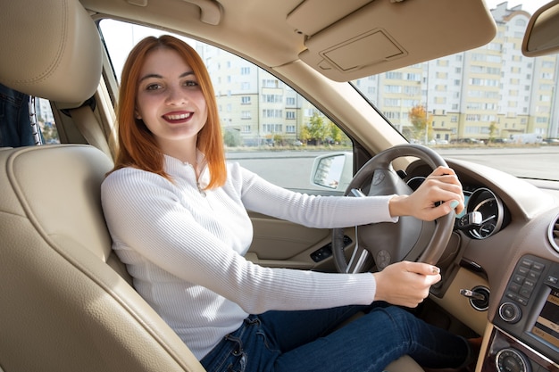 Giovane autista donna rossa dietro una ruota alla guida di un'auto sorridendo felicemente.