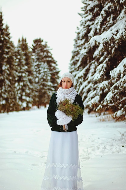 молодая рыжая женщина в зеленом свитере и белом свадебном платье гуляет в морозном зимнем парке.