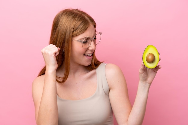 Young redhead woman holding an avocado isolated on pink background celebrating a victory