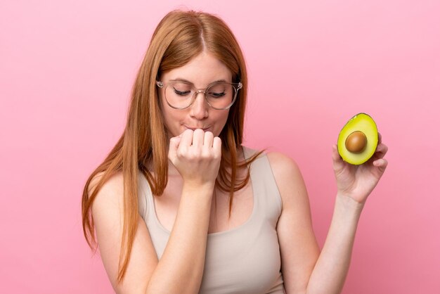 Young redhead woman holding an avocado isolated on pink background having doubts