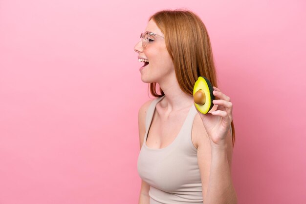 Young redhead woman holding an avocado isolated on pink background laughing in lateral position