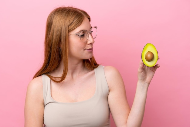 Young redhead woman holding an avocado isolated on pink background looking to the side