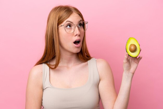 Young redhead woman holding an avocado isolated on pink background looking up and with surprised expression