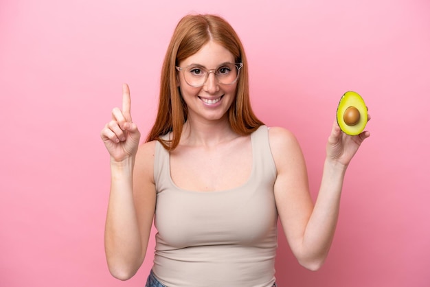 Young redhead woman holding an avocado isolated on pink background pointing up a great idea