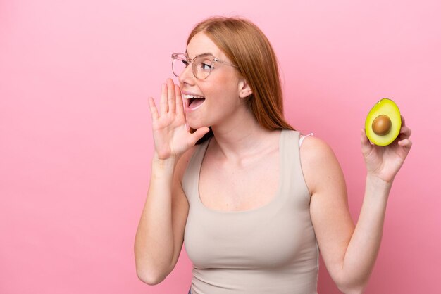 Young redhead woman holding an avocado isolated on pink\
background shouting with mouth wide open to the side