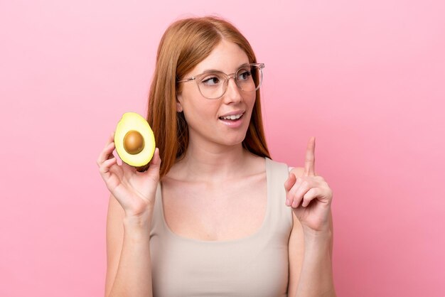 Young redhead woman holding an avocado isolated on pink background thinking an idea pointing the finger up