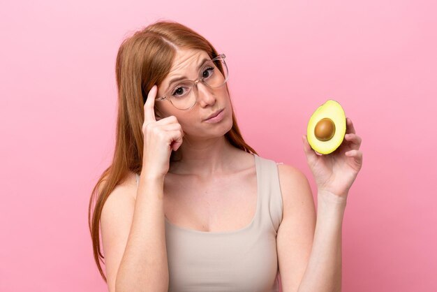 Young redhead woman holding an avocado isolated on pink background thinking an idea