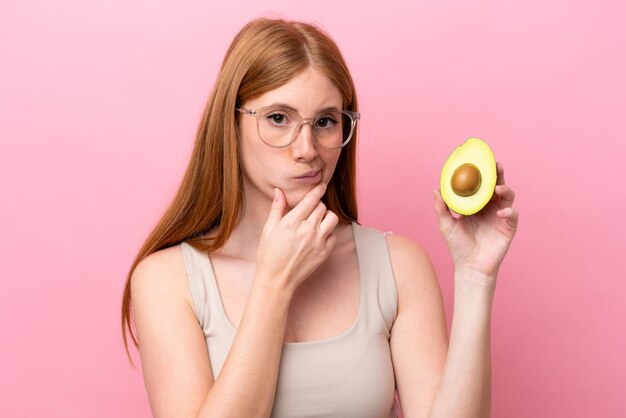 Young redhead woman holding an avocado isolated on pink background thinking