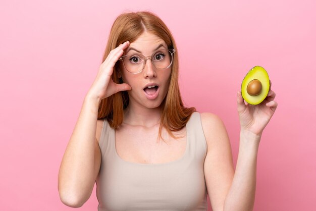 Young redhead woman holding an avocado isolated on pink background with surprise expression