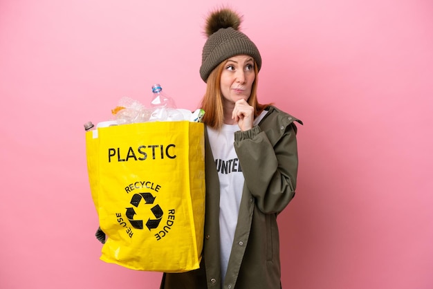 Young redhead woman holding a bag full of plastic bottles to recycle isolated on pink background having doubts and thinking