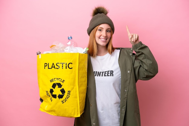 Young redhead woman holding a bag full of plastic bottles to recycle isolated on pink background pointing up a great idea