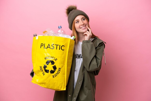 Young redhead woman holding a bag full of plastic bottles to recycle isolated on pink background thinking an idea while looking up