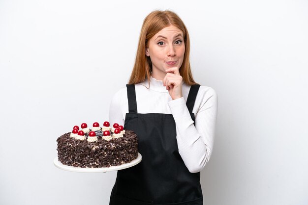 Young redhead woman holding birthday cake isolated on white background having doubts and thinking