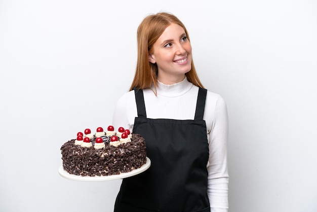 Young redhead woman holding birthday cake isolated on white background thinking an idea while looking up