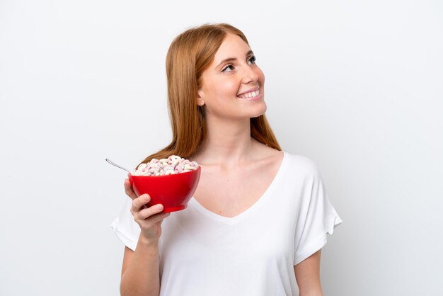 Young redhead woman holding a bowl of cereals isolated on white background looking up while smiling