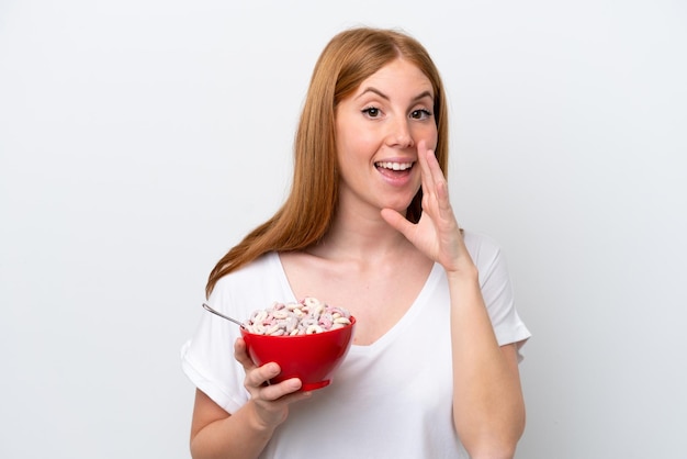 Young redhead woman holding a bowl of cereals isolated on white background shouting with mouth wide open