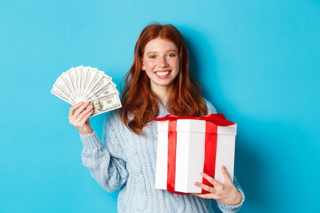 クリスマスのギフトボックスとお金を持って、幸せそうに笑って、青い背景の上に立っている若い赤毛の女性。