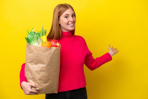 黄色の背景に食料品の買い物袋を持っている若い赤毛の女性が来るように誘うために手を側に伸ばす