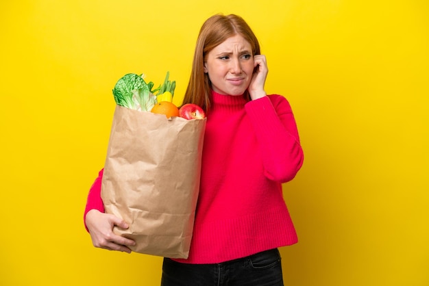 Young redhead woman holding a grocery shopping bag isolated on yellow background frustrated and covering ears