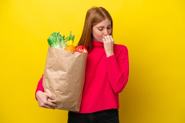 疑いを持って黄色の背景に分離された食料品の買い物袋を保持している若い赤毛の女性