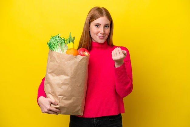 Young redhead woman holding a grocery shopping bag isolated on yellow background inviting to come with hand happy that you came