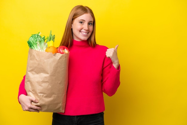 Young redhead woman holding a grocery shopping bag isolated on yellow background pointing to the side to present a product