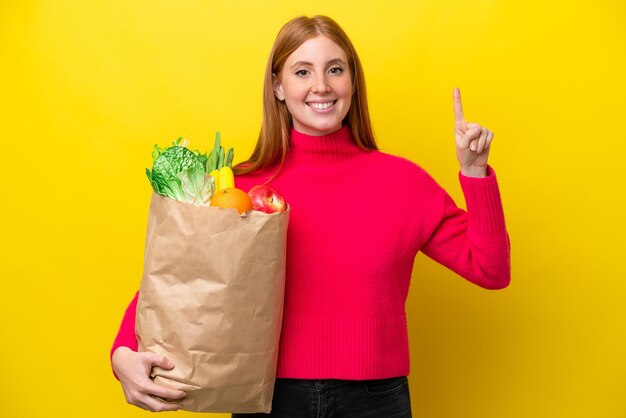 素晴らしいアイデアを指している黄色の背景に分離された食料品の買い物袋を保持している若い赤毛の女性