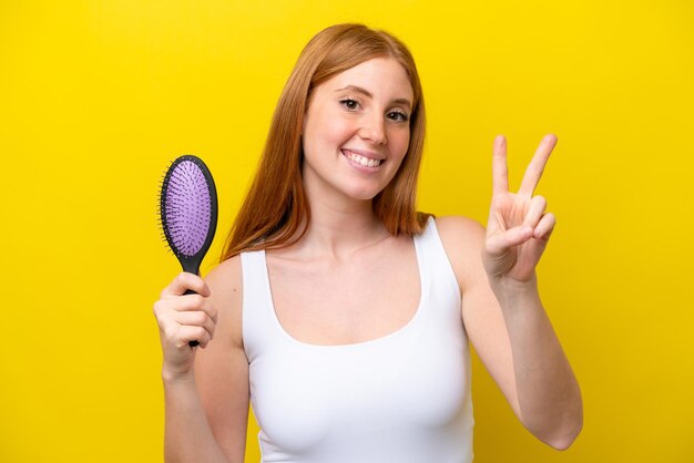 笑顔で勝利のサインを示す白い背景で隔離のヘアブラシを保持している若い赤毛の女性