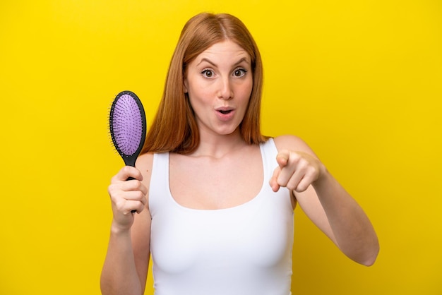 Young redhead woman holding a hairbrush isolated on white background surprised and pointing front
