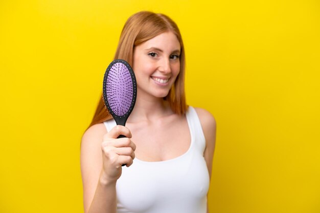 Giovane donna rossa che tiene una spazzola per capelli isolata su sfondo bianco con un'espressione felice