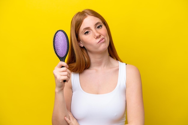 悲しい表情で白い背景で隔離のヘアブラシを保持している若い赤毛の女性