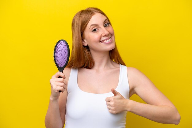 何か良いことが起こったので、親指で白い背景に分離されたヘアブラシを保持している若い赤毛の女性