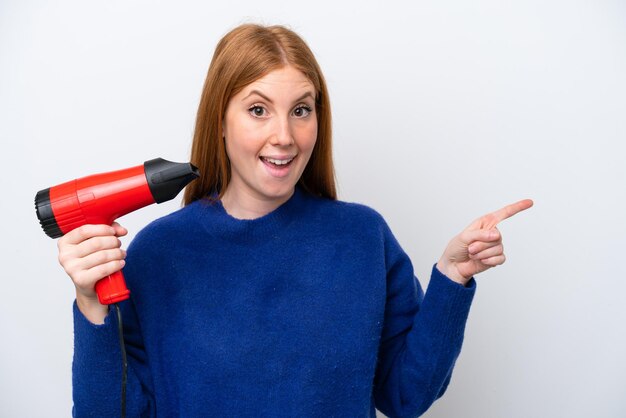 Young redhead woman holding a hairdryer isolated on white background surprised and pointing finger to the side