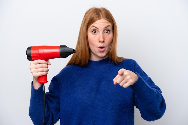 Young redhead woman holding a hairdryer isolated on white background surprised and pointing front