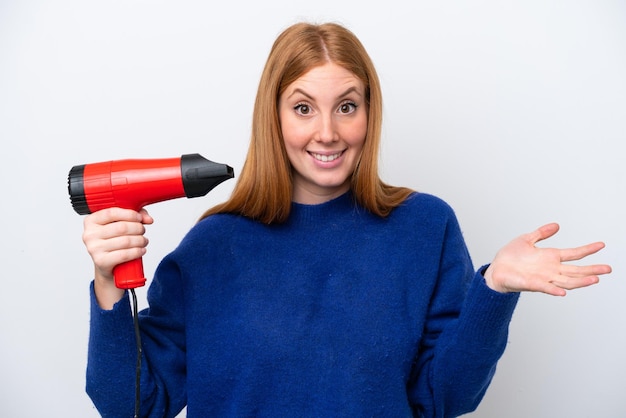 Young redhead woman holding a hairdryer isolated on white background with shocked facial expression